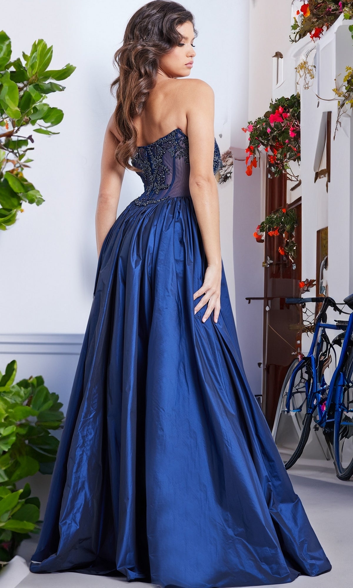 A woman stands outdoors near lush plants and a blue bicycle, wearing the Jovani Strapless Long Taffeta Prom Dress 26114 featuring a beaded sheer bodice and full skirt.
